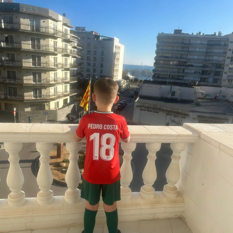 Child wearing a red sports jersey with 'Pedro Costa 18' on a balcony overlooking buildings and a flag.