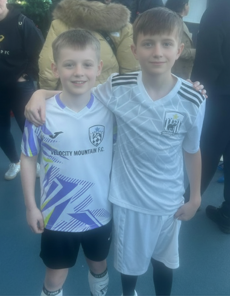 Two young boys in soccer uniforms standing together with a crowd in the background.